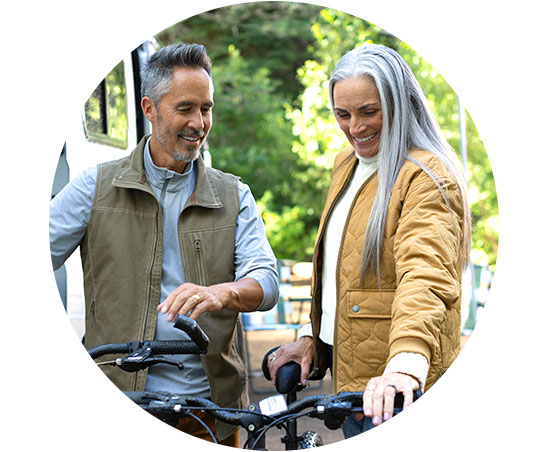 Twee personen, een man en een vrouw, staan naast fietsen en lijken een gesprek te voeren. Ze bevinden zich buiten in een groene, zonnige omgeving. Geen tekst zichtbaar.