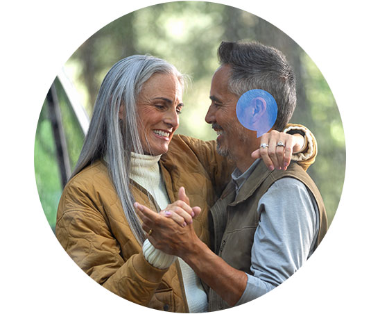 Een oudere man met een zichtbaar blauw hoortoestel danst met een vrouw in een casual buitenomgeving omringd door groen. Hun gezichten zijn geblurd, en ze lijken gelukkig.