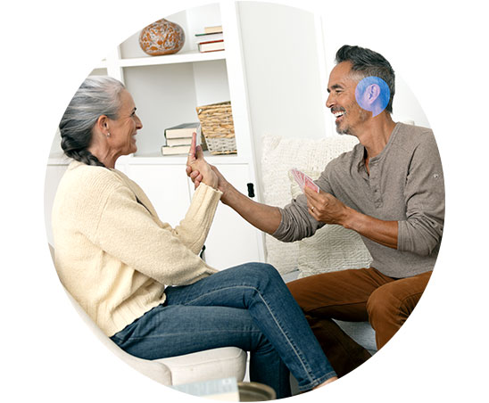 Een oudere vrouw en man zitten in een huiskamer op stoelen en lijken een kaartspel te spelen. Op de achtergrond zijn boekenplanken met decoratieve items zichtbaar.